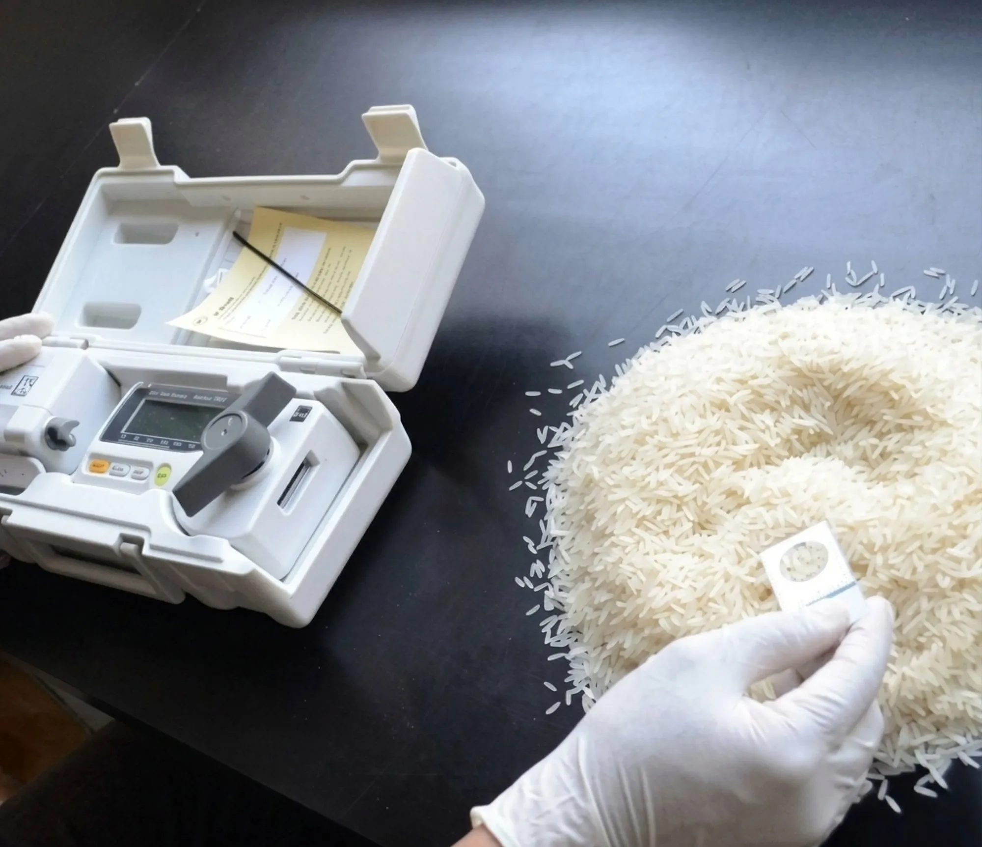 A portable grain moisture meter and its sample cell beside a heap of milled Rice